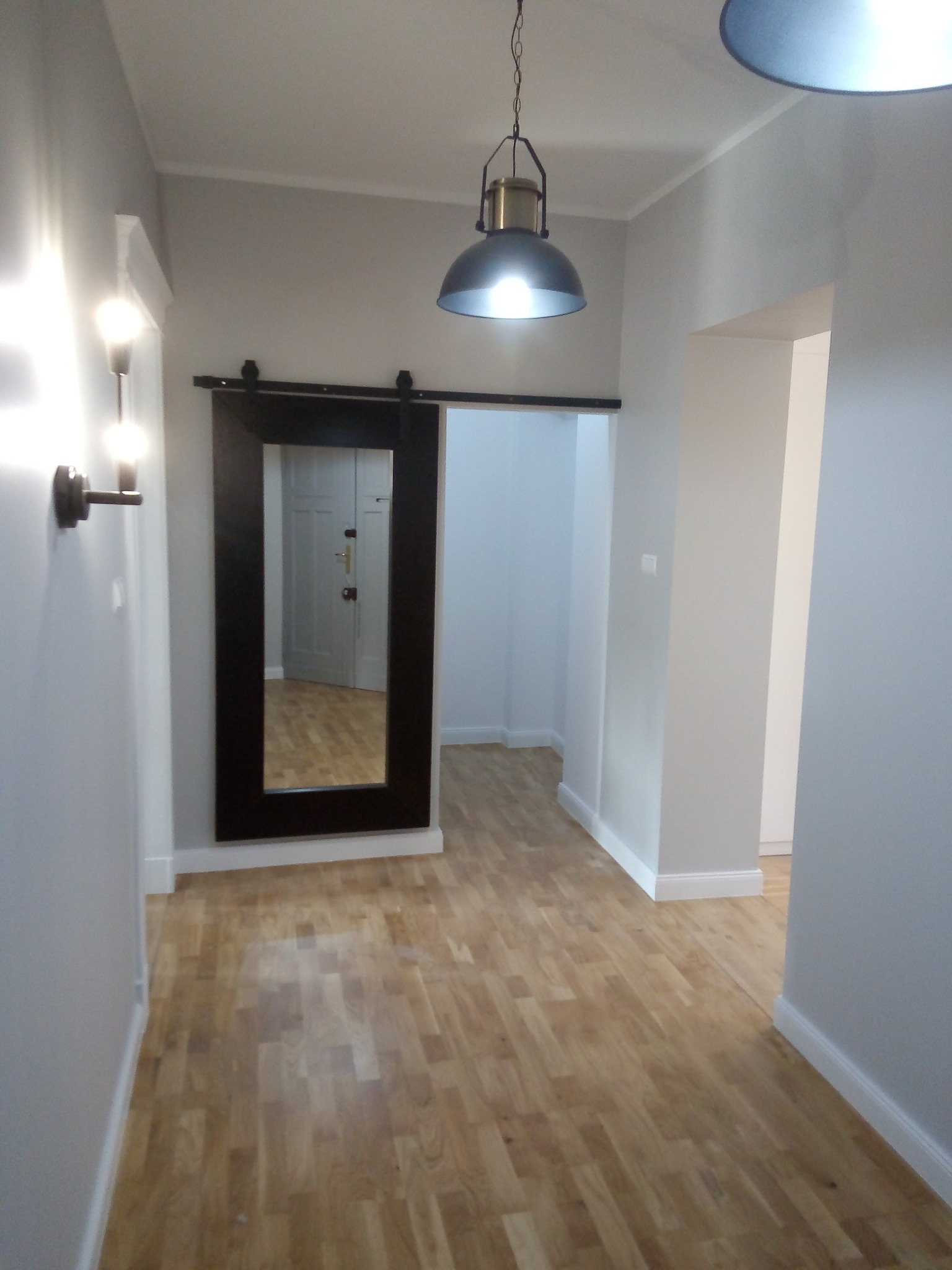 Wnętrze korytarza z drewnianą podłogą, przesuwne lustro w czarnej ramie, lampy wiszące w stylu industrialnym i kinkiet na ścianie.