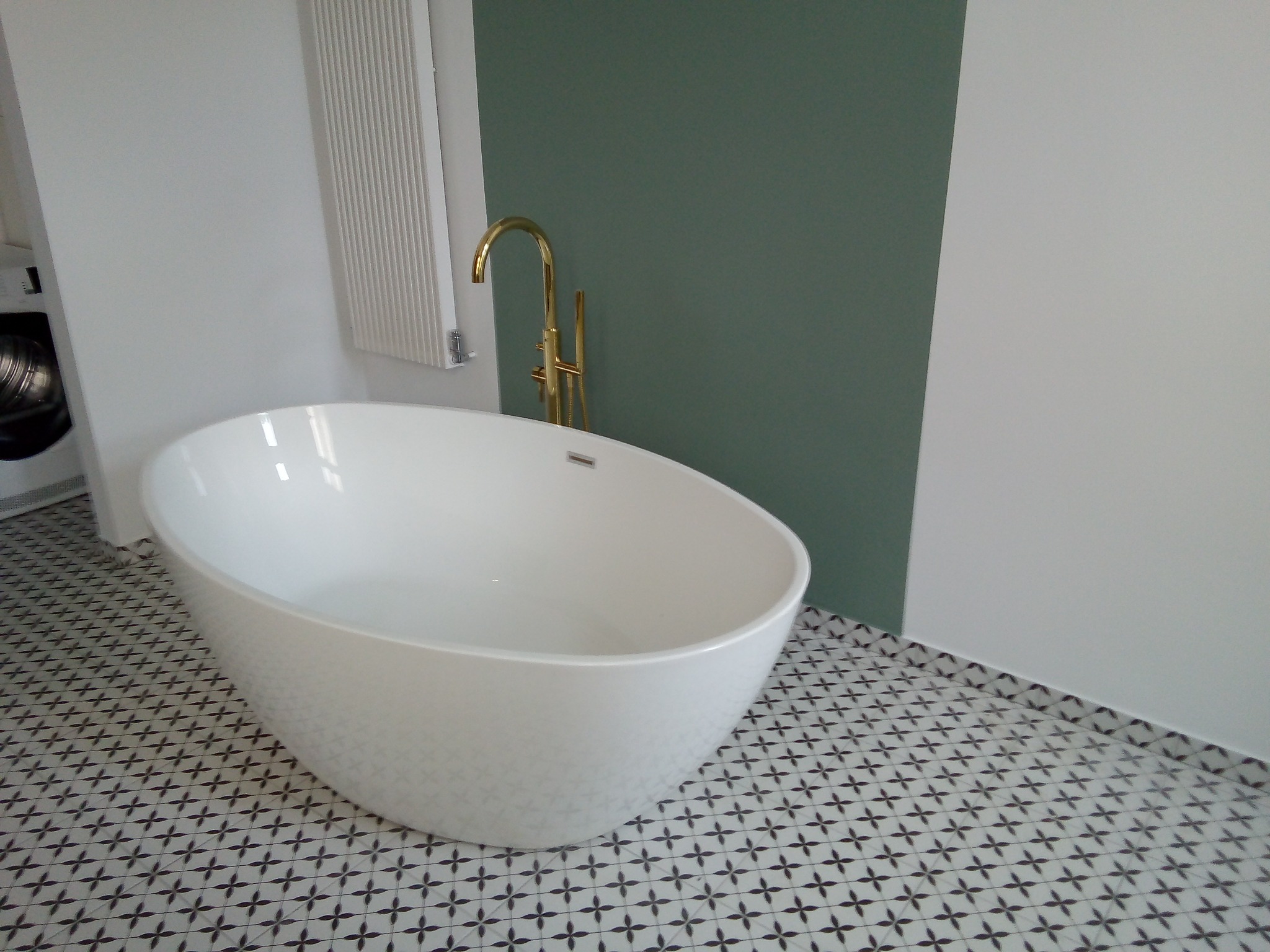 Wolnostojąca wanna o owalnym kształcie z błyszczącą, złotą armaturą na tle ściany pomalowanej na zielono i biało, oraz podłogi wyłożonej płytkami w geometryczny wzór.