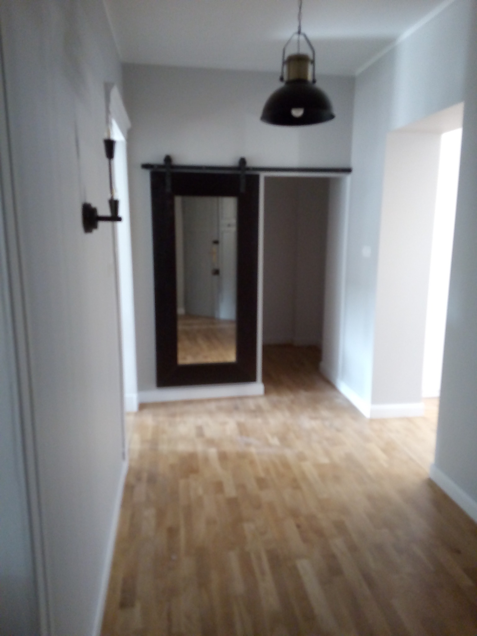 Wnętrze mieszkania po remoncie: drewniana podłoga, przesuwne drzwi w ciemnej ramie z widokiem na kolejne pomieszczenie, szara ściana z kinkietem i wisząca lampa w stylu industrialnym.
