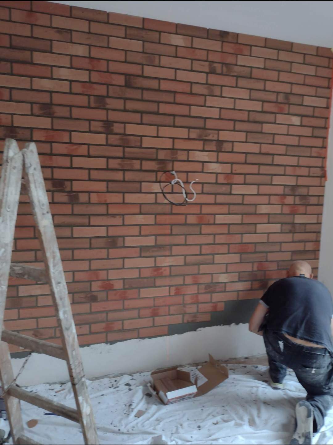 Montaż okładziny ściennej imitującej cegłę, widoczny fragment ściany z niedokończonym fragmentem, drabina malarska i instalacja elektryczna ze zwisającymi kablami.