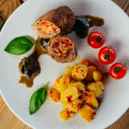 Elegancko podane roladki wołowe przekrojone na pół, ziemniaki z przyprawami, pieczone śliwki, pomidorki koktajlowe, gałązka rozmarynu i liście bazylii na białym talerzu z fakturą, na drewnianym stole.