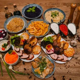 Bogato zastawiony drewniany stół z różnorodnymi daniami: pieczone mięsa, ryby, frytki, surówki, puree, sosy w metalowych naczyniach, buteleczki z olejem i octem, przyprawy i świeży czosnek.