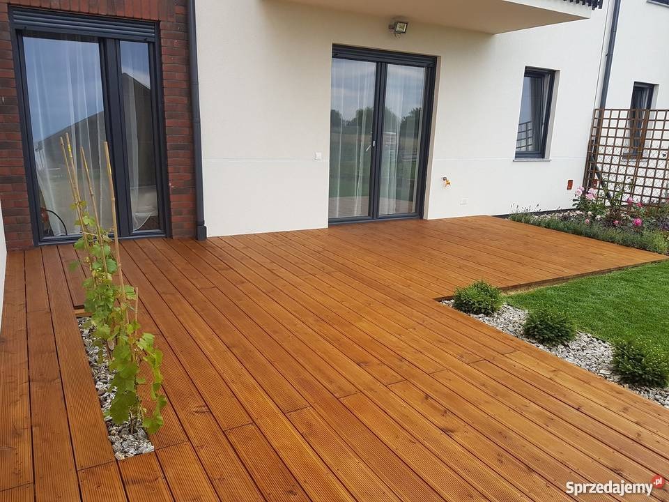 Drewniany taras przydomowy z desek w ciepłym odcieniu brązu, z wbudowaną donicą i roślinami pnącymi, obok trawnik z ozdobnymi krzewami i kratką na pnącza.