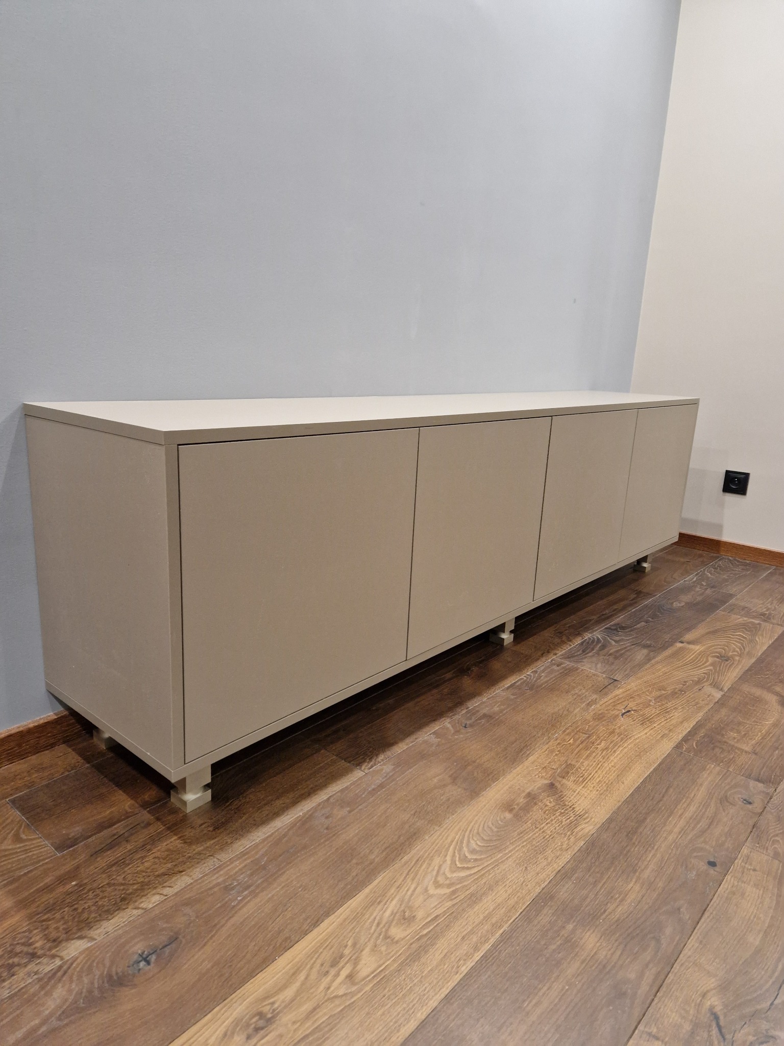 Beżowa szafka RTV na niskich nóżkach, ustawiona na drewnianej podłodze, na tle szarej ściany.