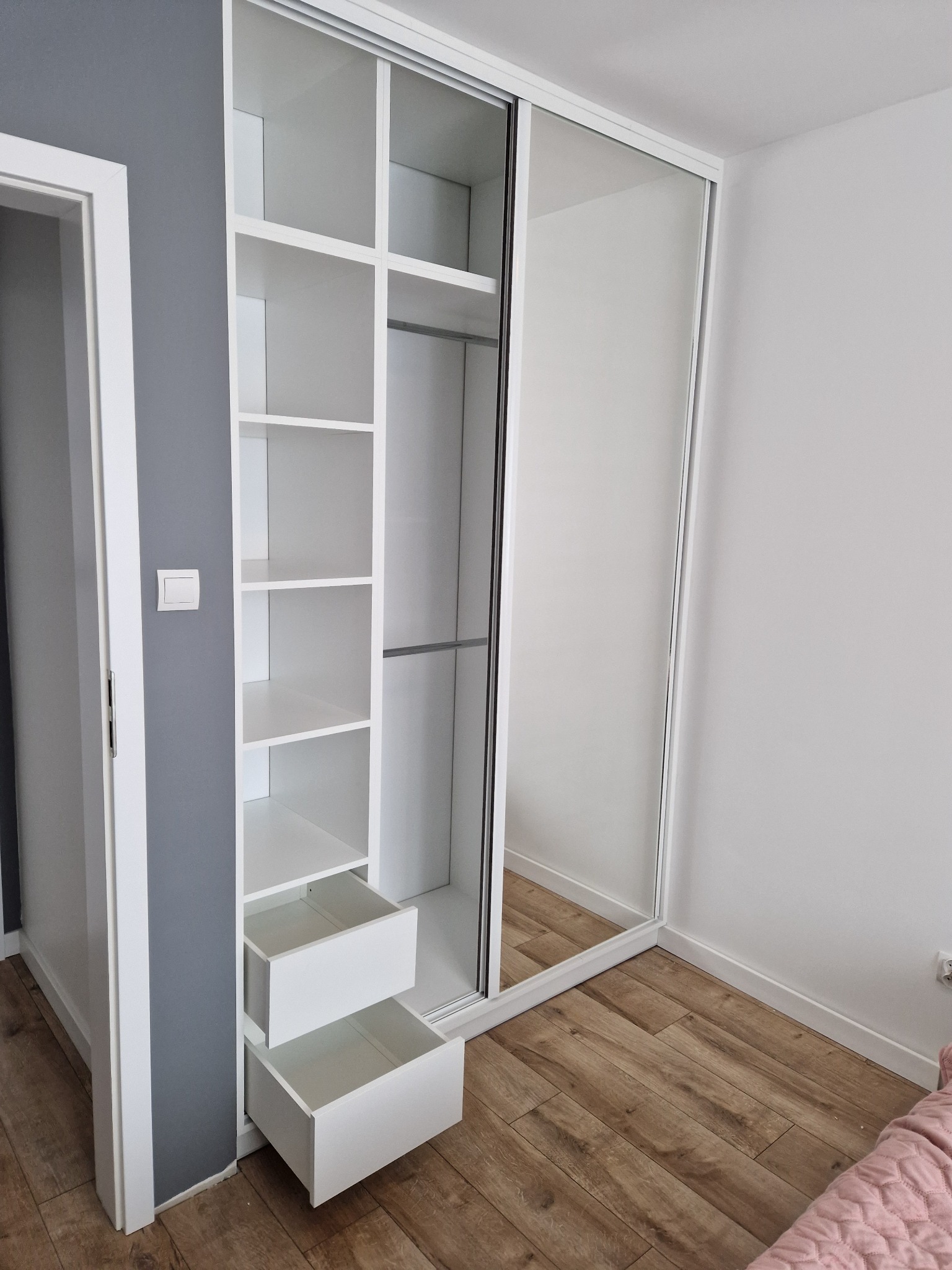 Biała szafa wnękowa z przesuwnymi drzwiami lustrzanymi i otwartymi półkami z szufladami, na drewnianej podłodze, widok z boku.