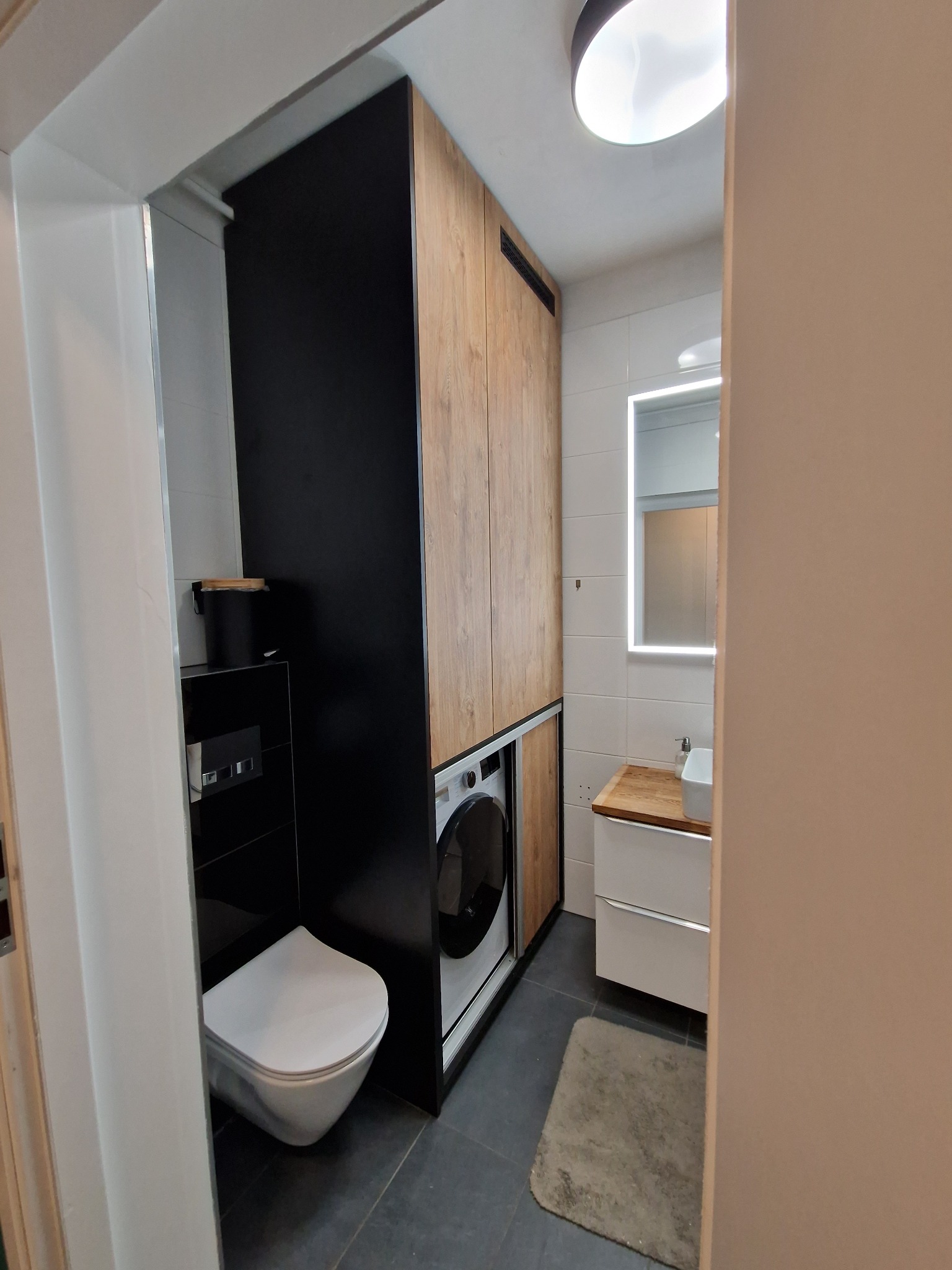 Nowoczesna łazienka w minimalistycznym stylu z zabudowaną pralką w szafie z drewnianymi frontami, wisząca toaleta, umywalka z drewnianym blatem i podświetlane lustro.