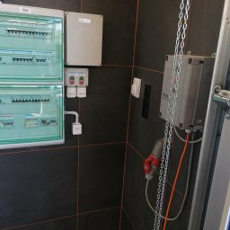 IPINSTAL Usługi Elektryczne Paweł Świstek - Rozdzielnica elektryczna z zieloną obudową na ciemnej ścianie z pomarańczowymi fugami, obok skrzynka sterująca zasilana kablem z czerwoną wtyczką, łańcuch i mechanizm bramy garażowej.