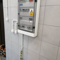 IPINSTAL Usługi Elektryczne Paweł Świstek - Zamknięta, biała skrzynka rozdzielcza na kafelkowanej ścianie z widocznymi bezpiecznikami, wyłącznikiem głównym i naklejką ostrzegawczą 'Nie dotykać urządzenia elektrycznego', z widocznym przewodem...