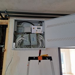 IPINSTAL Usługi Elektryczne Paweł Świstek - Otwarta skrzynka z rozdzielnicą kablową, widoczne kable koncentryczne i urządzenia, drabina z narzędziami w pomieszczeniu w trakcie prac instalacyjnych.