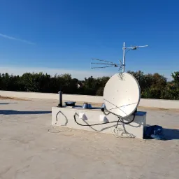 Antena satelitarna i antena naziemna zamontowane na betonowym dachu budynku, widoczne kable i elementy montażowe, na tle błękitne niebo i zieleń drzew.