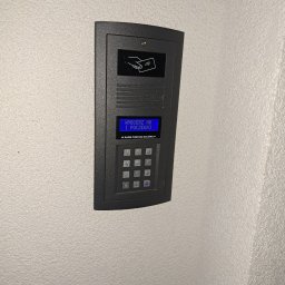 IPINSTAL Usługi Elektryczne Paweł Świstek - Szary panel domofonu z czytnikiem kart i klawiaturą numeryczną, zamontowany na jasnej, teksturowanej ścianie, z niebieskim wyświetlaczem instrukcji w języku polskim.