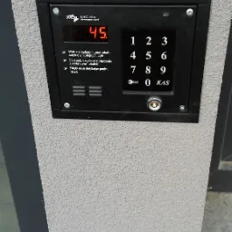 Czarny panel domofonu z wyświetlaczem LED na szarej, teksturowanej ścianie, z przyciskami numerycznymi i instrukcją obsługi.