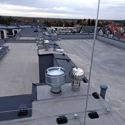 IPINSTAL Usługi Elektryczne Paweł Świstek - Płaski dach budynku z zainstalowaną instalacją odgromową, widoczne przewody, uchwyty i maszt odgromowy na tle zabudowy mieszkalnej w pochmurny dzień.