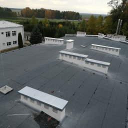 IPINSTAL Usługi Elektryczne Paweł Świstek - Widok z góry na płaski dach budynku z szarą papą, kominami wentylacyjnymi i zainstalowanym systemem odgromowym z metalowymi masztami i przewodami.