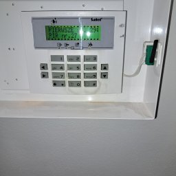 IPINSTAL Usługi Elektryczne Paweł Świstek - Panel sterowania systemu alarmowego firmy Satel, zamontowany na ścianie, z wyświetlaczem pokazującym komunikat 'PIERWSZE PIR nr.27', widoczne przyciski numeryczne i funkcyjne, całość w białej...