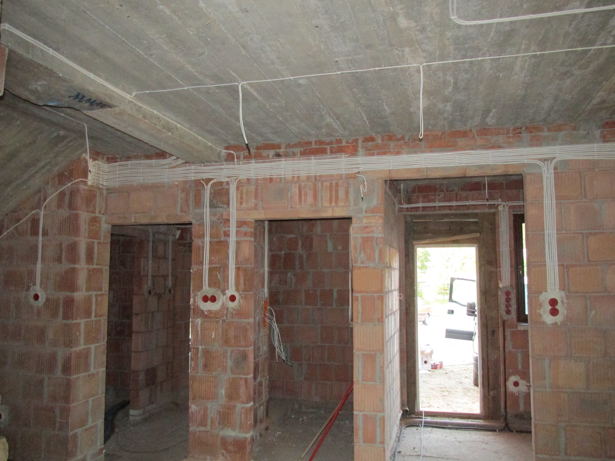 Surowy stan wnętrza z cegły, widoczne białe kable elektryczne prowadzone po suficie i ścianach, puszki instalacyjne z czerwonymi zaślepkami, okablowanie w trakcie prac.