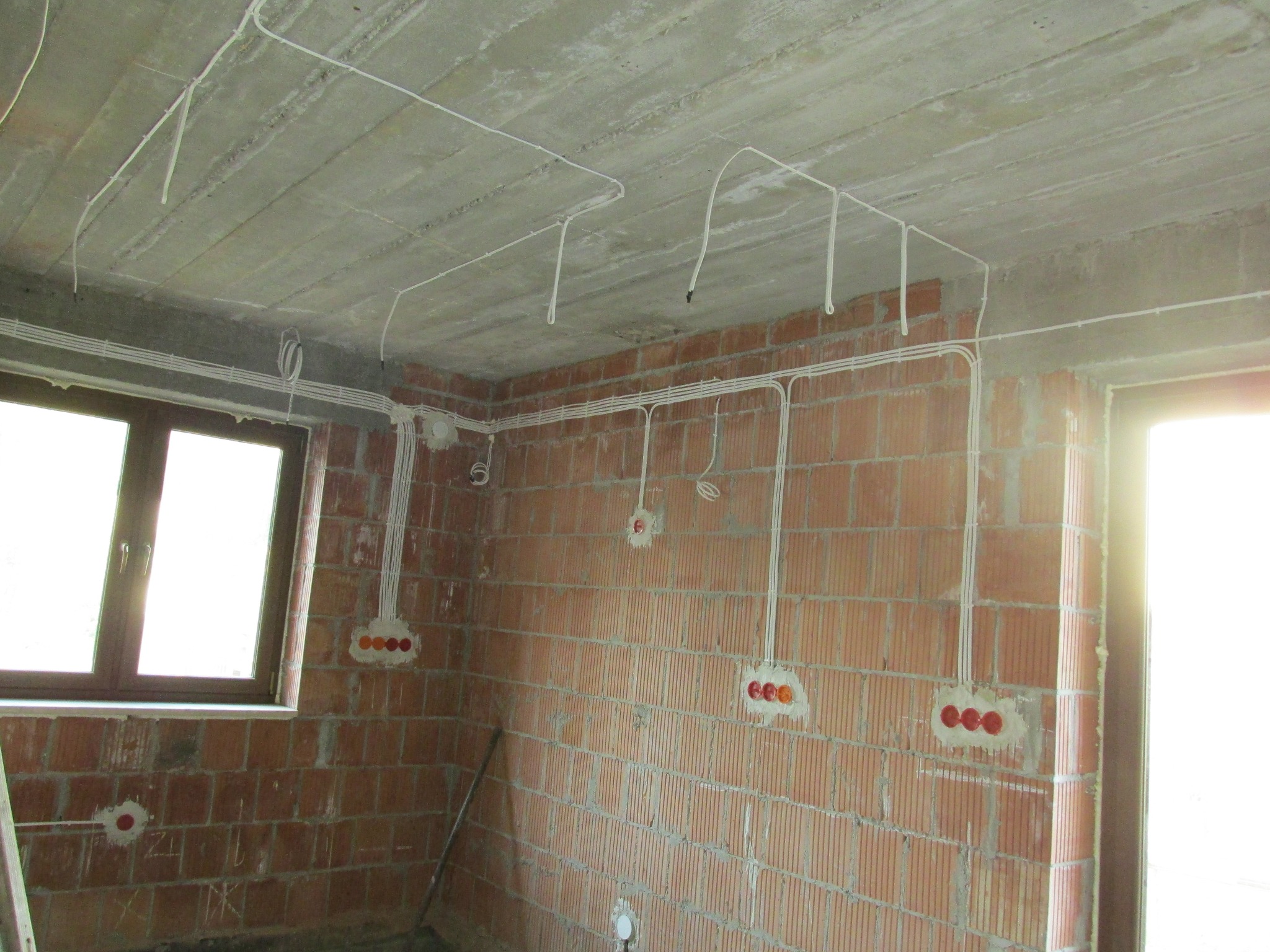 Surowy stan: przewody elektryczne rozprowadzone po ścianach z czerwonej cegły i pod betonowym sufitem w nowym budynku, widoczne puszki instalacyjne.