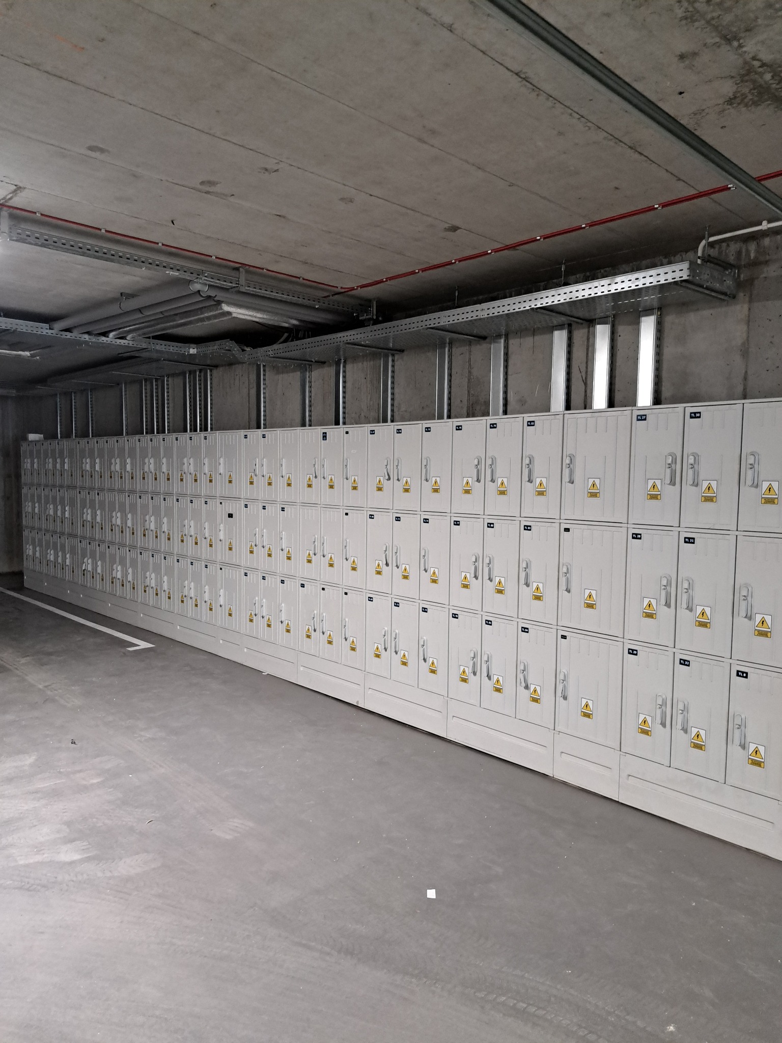 Rząd szarych, metalowych szafek elektrycznych z żółtymi znakami ostrzegawczymi, umieszczonych w surowym, betonowym pomieszczeniu z widocznymi kanałami kablowymi pod sufitem.