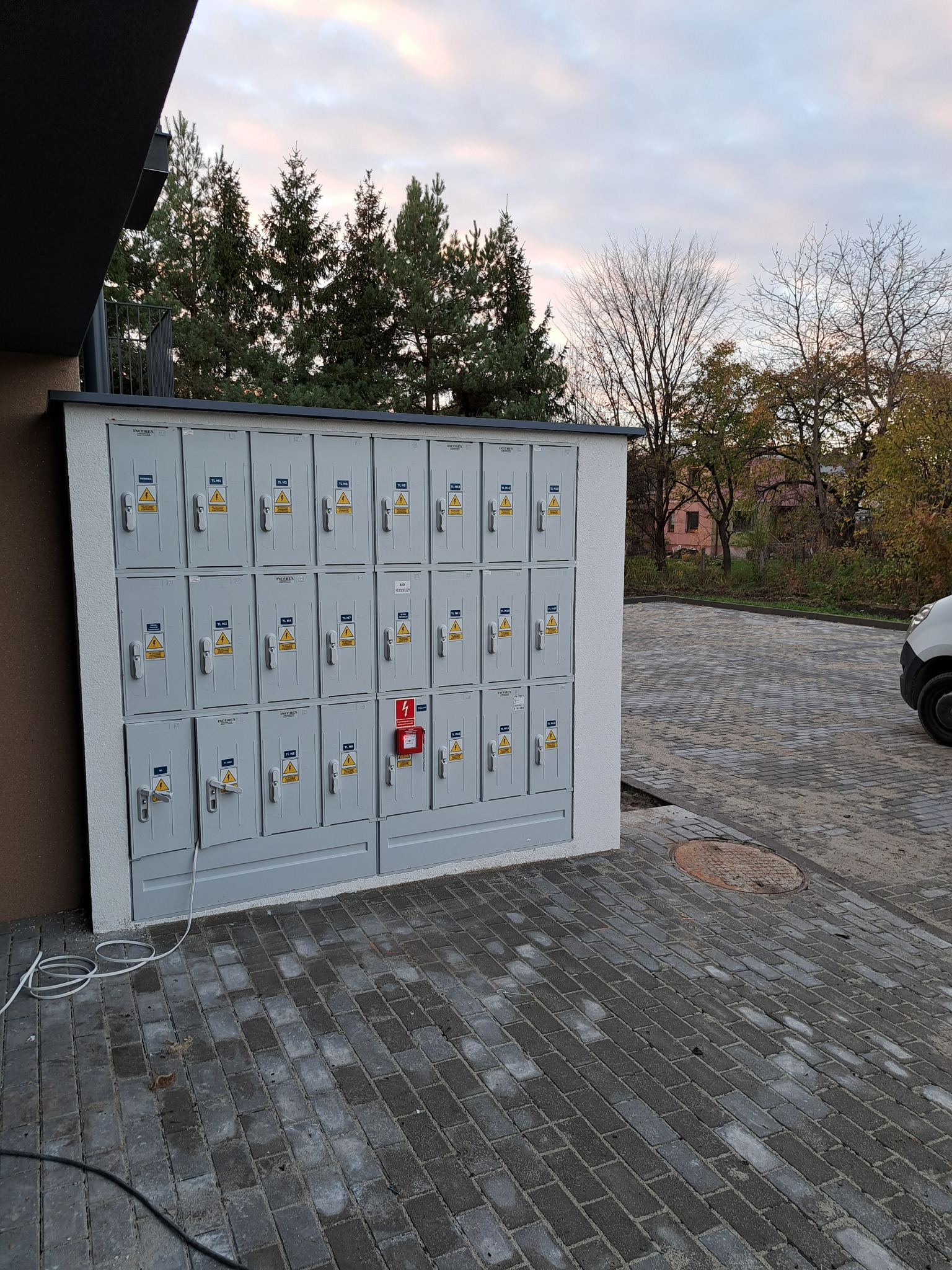 Zewnętrzna szafa rozdzielcza z wieloma zabezpieczonymi skrzynkami, umieszczona na nowym osiedlu z kostki brukowej, widoczny przewód zasilający.