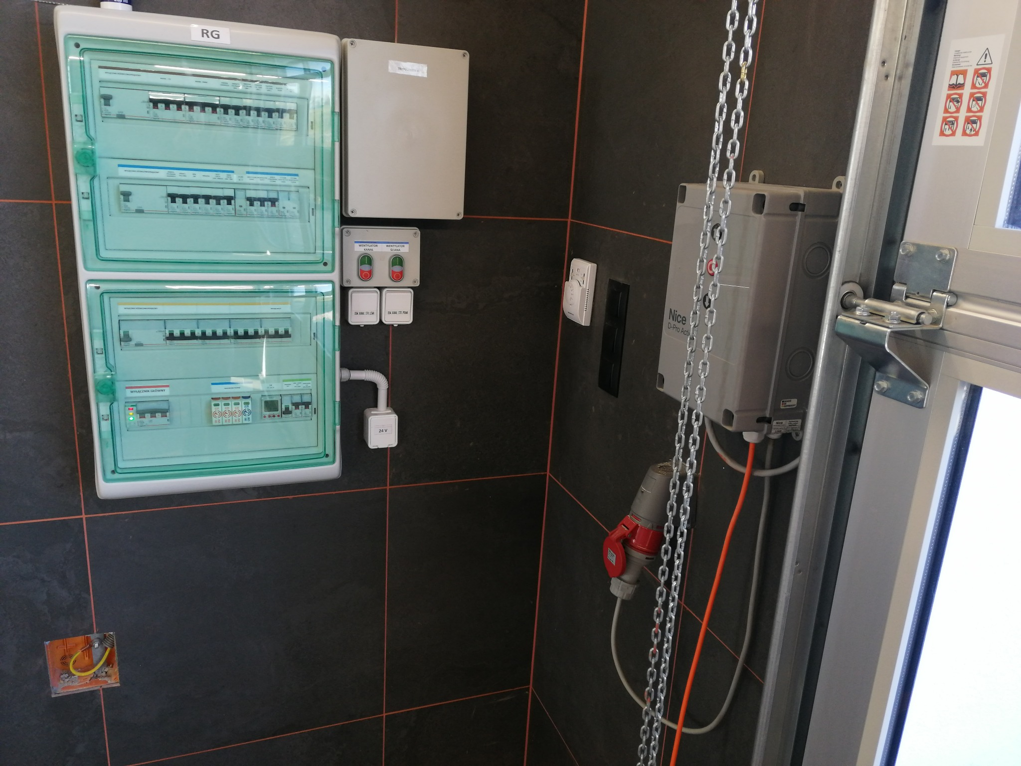 Rozdzielnica elektryczna z zieloną obudową na ciemnej ścianie z pomarańczowymi fugami, obok skrzynka sterująca zasilana kablem z czerwoną wtyczką, łańcuch i mechanizm bramy garażowej.