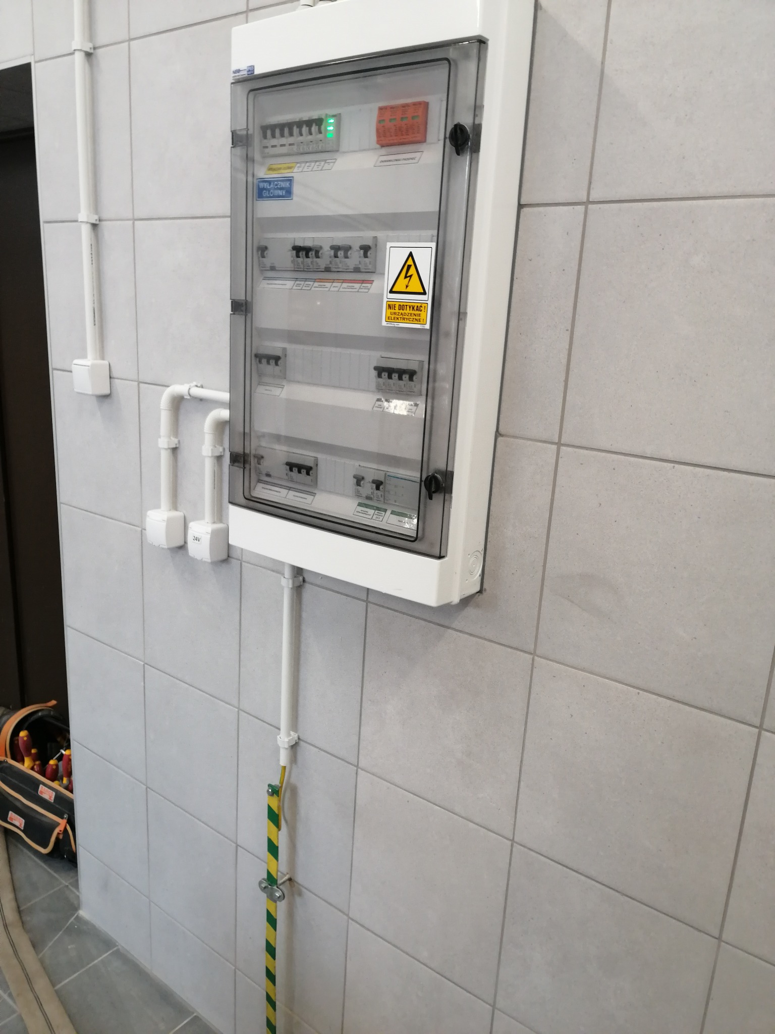Zamknięta, biała skrzynka rozdzielcza na kafelkowanej ścianie z widocznymi bezpiecznikami, wyłącznikiem głównym i naklejką ostrzegawczą 'Nie dotykać urządzenia elektrycznego', z widocznym przewodem...