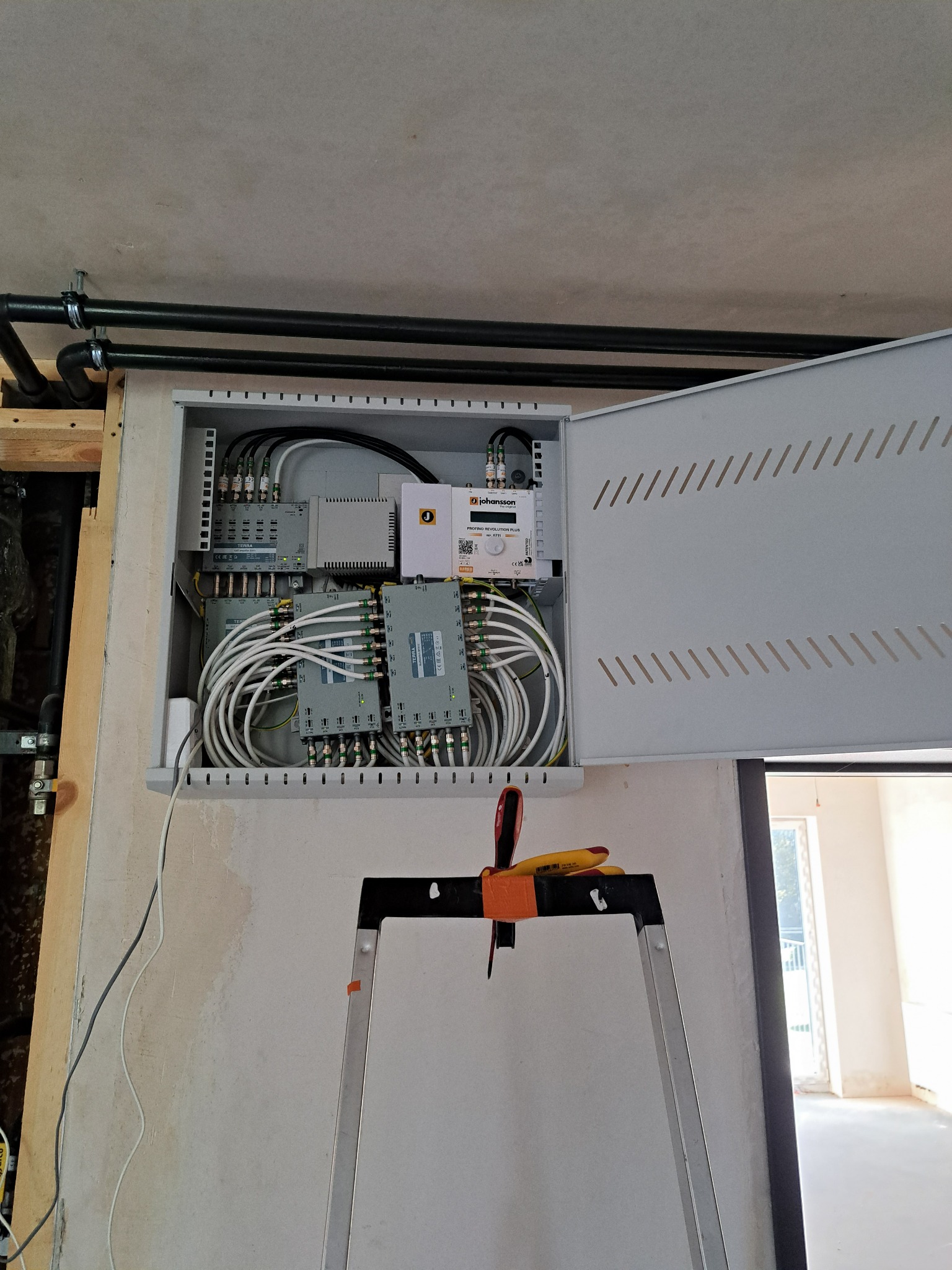 Otwarta skrzynka z rozdzielnicą kablową, widoczne kable koncentryczne i urządzenia, drabina z narzędziami w pomieszczeniu w trakcie prac instalacyjnych.