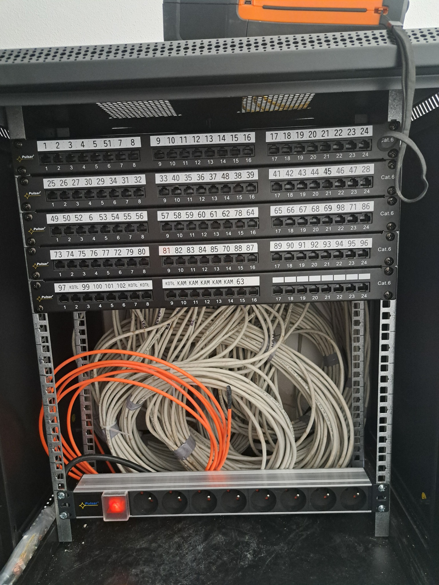 Panel krosowniczy z podłączonymi kablami sieciowymi, widoczny również przedłużacz z czerwonym włącznikiem w szafie rack.
