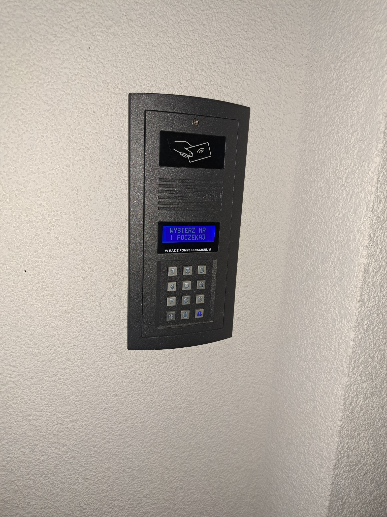 Szary panel domofonu z czytnikiem kart i klawiaturą numeryczną, zamontowany na jasnej, teksturowanej ścianie, z niebieskim wyświetlaczem instrukcji w języku polskim.
