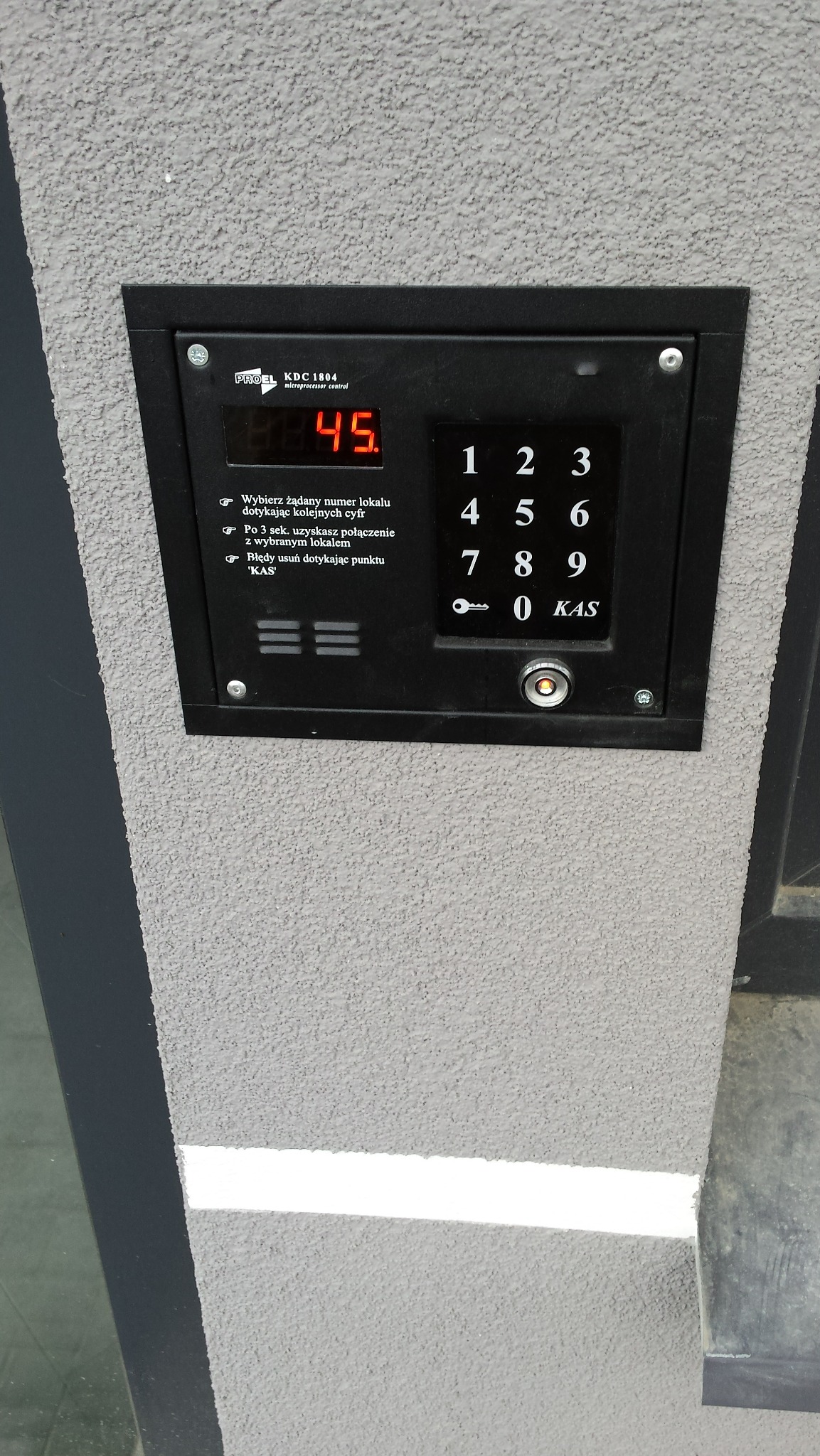 Czarny panel domofonu z wyświetlaczem LED na szarej, teksturowanej ścianie, z przyciskami numerycznymi i instrukcją obsługi.