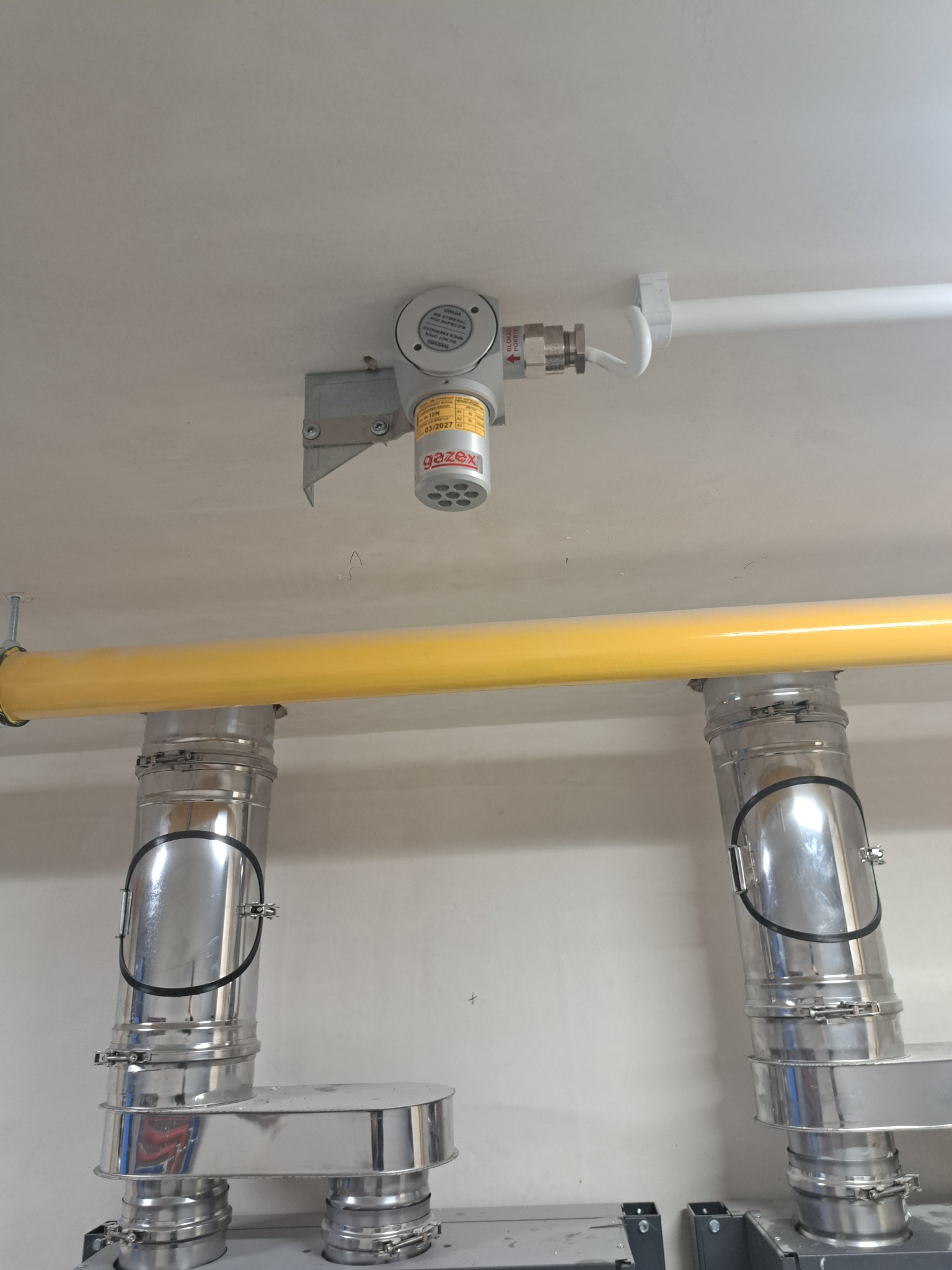 Zamontowany detektor gazu Gazex z żółtą rurą gazową oraz dwoma srebrnymi rurami odprowadzającymi spaliny pod sufitem.