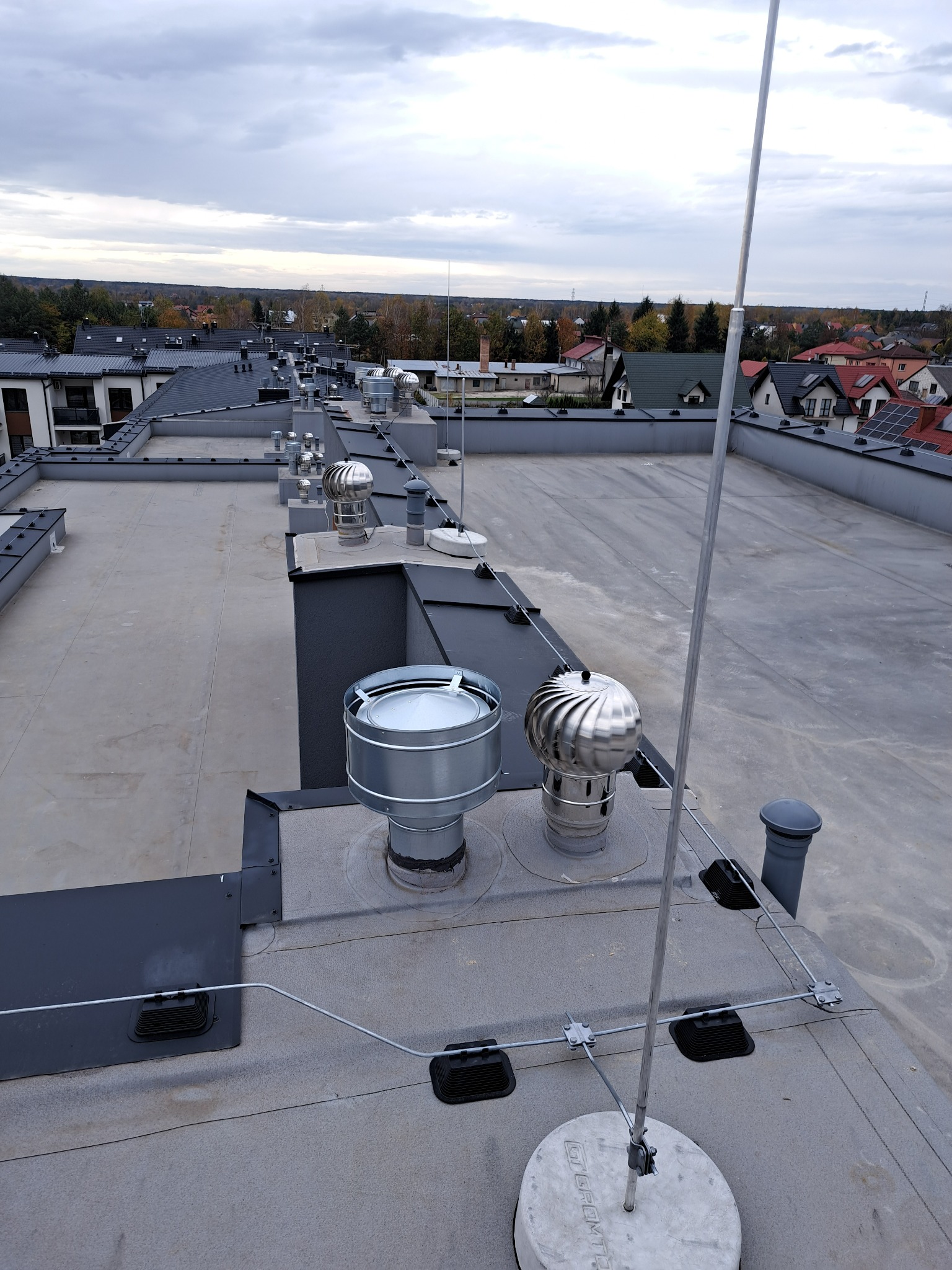 Płaski dach budynku z zainstalowaną instalacją odgromową, widoczne przewody, uchwyty i maszt odgromowy na tle zabudowy mieszkalnej w pochmurny dzień.