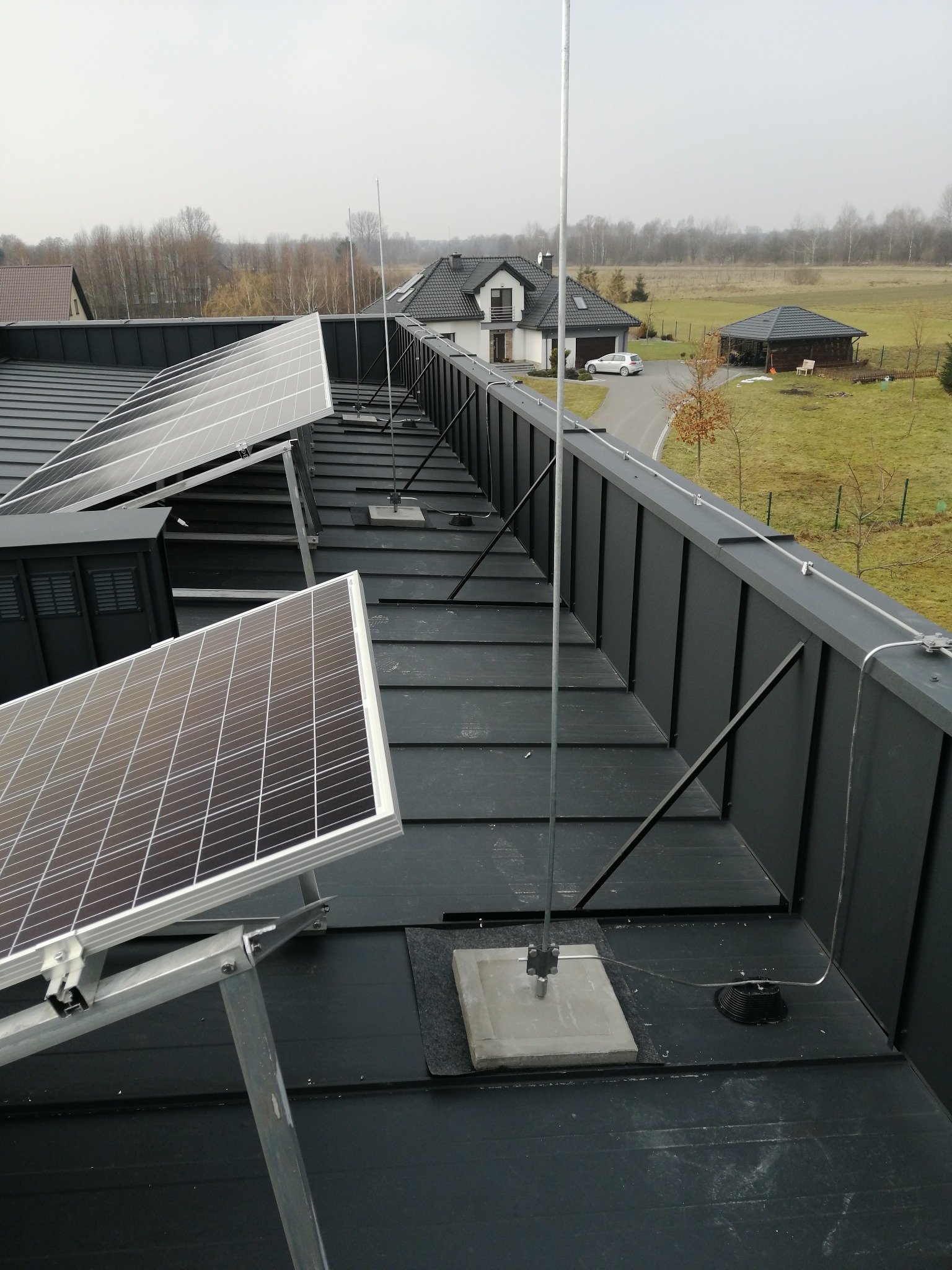 Widok z góry na instalację paneli słonecznych i masztów odgromowych na dachu domu jednorodzinnego w pochmurny dzień.