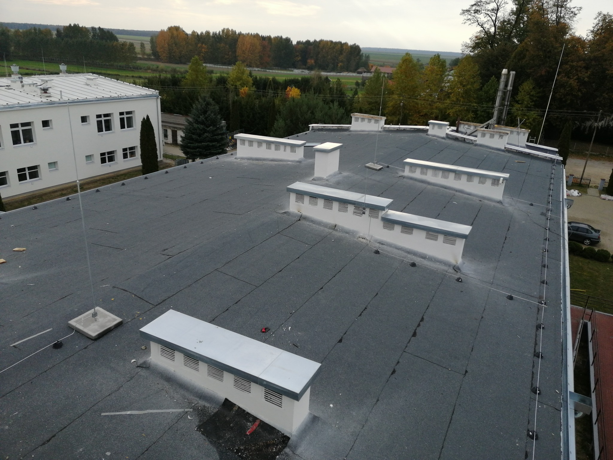 Widok z góry na płaski dach budynku z szarą papą, kominami wentylacyjnymi i zainstalowanym systemem odgromowym z metalowymi masztami i przewodami.
