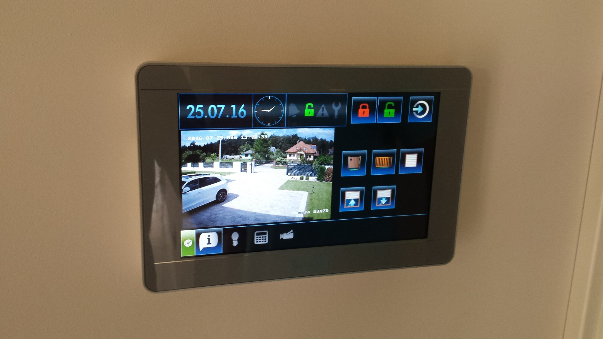 Ekran dotykowy systemu inteligentnego domu, wyświetlający podgląd z kamery monitoringu posesji z widocznym samochodem, bramą i budynkiem mieszkalnym.