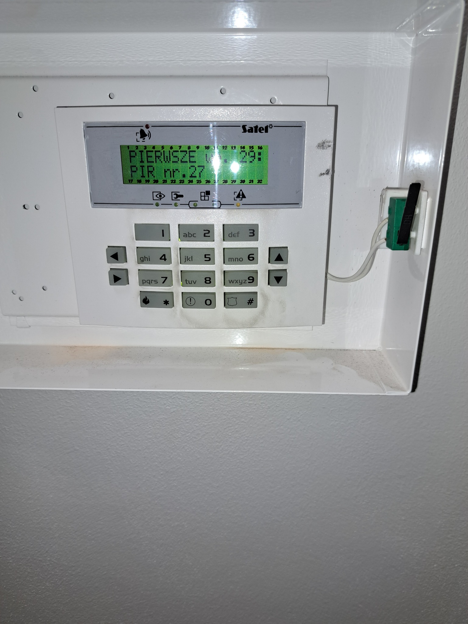 Panel sterowania systemu alarmowego firmy Satel, zamontowany na ścianie, z wyświetlaczem pokazującym komunikat 'PIERWSZE PIR nr.27', widoczne przyciski numeryczne i funkcyjne, całość w białej...