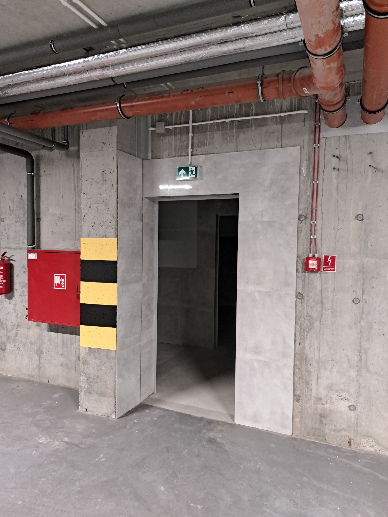 Wnętrze budynku w stanie surowym z widocznymi instalacjami pod sufitem, czerwona szafka hydrantowa i tablica ostrzegawcza obok wejścia ewakuacyjnego.
