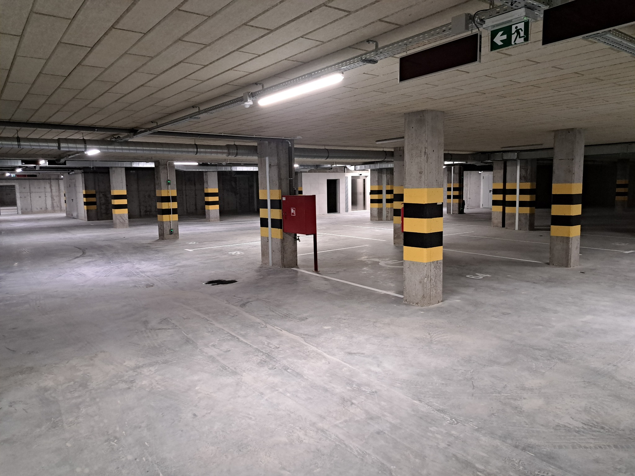 Oświetlony garaż podziemny z betonowymi kolumnami oznaczonymi czarno-żółtymi pasami, widoczne białe linie wyznaczające miejsca parkingowe, instalacje pod sufitem i znak ewakuacyjny.