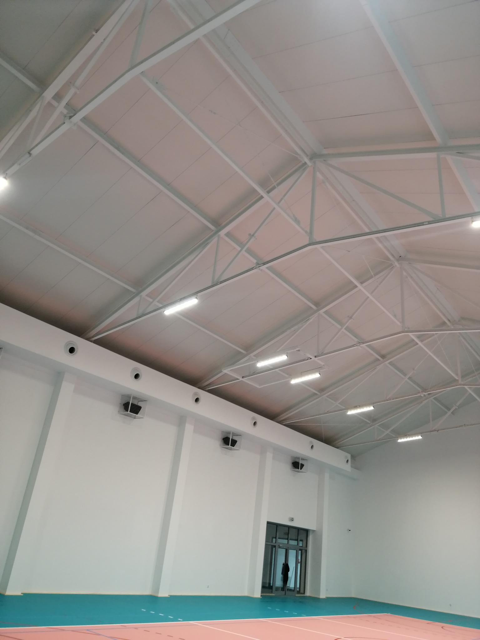 Wnętrze hali sportowej z widoczną konstrukcją dachu, oświetleniem liniowym LED i systemem wentylacji pod stropem, osoba w drzwiach w oddali.
