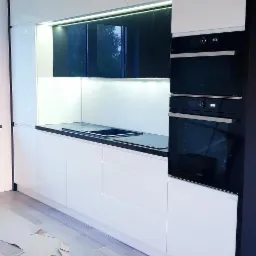 Nowoczesna, biała kuchnia z czarnymi akcentami, oświetleniem LED pod szafkami wiszącymi i dwoma piekarnikami w zabudowie, częściowo zabezpieczona podłoga podczas montażu.