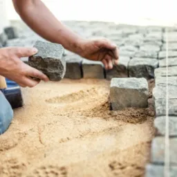 Pracownik w niebieskich spodniach umieszcza szarą kostkę brukową w piaskowym podsypie, obok leży młotek brukarski, widoczny fragment ułożonej nawierzchni.