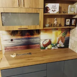 Fragment zabudowy kuchennej z szafkami z drewnopodobnym fornirem, szarymi frontami dolnych szafek i szkłem dekoracyjnym z motywem tropikalnym na ścianie między blatem a szafkami wiszącymi;...