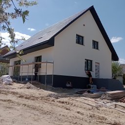 Pol-welder - Dwukondygnacyjny dom z czarnym dachem i białymi ścianami podczas prac wykończeniowych elewacji; dwóch pracowników na rusztowaniach maluje dolną część budynku na szaro, a w tle kwitnące drzewo owocowe.