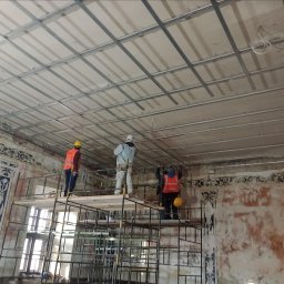 Pol-welder - Trzech pracowników na rusztowaniach montuje metalowy stelaż pod sufit podwieszany w pomieszczeniu z dekoracyjnymi, ale zniszczonymi ścianami.