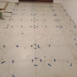 Pol-welder - Układanie jasnych płytek podłogowych z systemem poziomowania, widoczne niebieskie kliny i klipsy dystansowe.