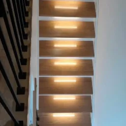 Drewniane schody z wbudowanym oświetleniem LED, czarne metalowe balustrady po lewej stronie, białe ściany.