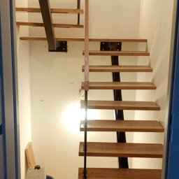 Pol-welder - Nowoczesne schody drewniane z metalowymi elementami, widoczne z dołu. Podłoga wyłożona parkietem w jodełkę. W tle białe ściany i widoczne narzędzia.