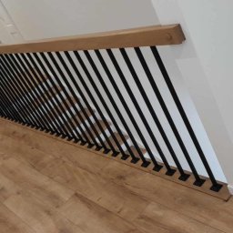 Pol-welder - Nowoczesne schody z drewnianą poręczą i czarnymi, pionowymi balustradami, widok z góry na parkietową podłogę. Minimalistyczny design, jasne ściany, wykończenie wnętrza.