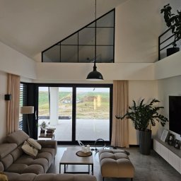 Pol-welder - Jasne wnętrze salonu z dużym oknem na ogród. Minimalistyczny design, beżowa sofa, drewniany stolik kawowy, wysokie okno trójkątne. Rośliny doniczkowe dodają naturalnego akcentu.