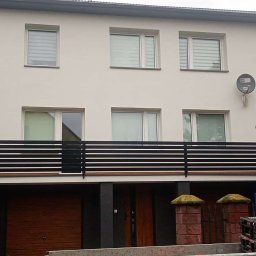 Pol-welder - Dwukondygnacyjny dom z jasną elewacją, czarną balustradą balkonu i numerem 6. Widok z poziomu ulicy, częściowo zasłonięty przez roślinność i samochód dostawczy.