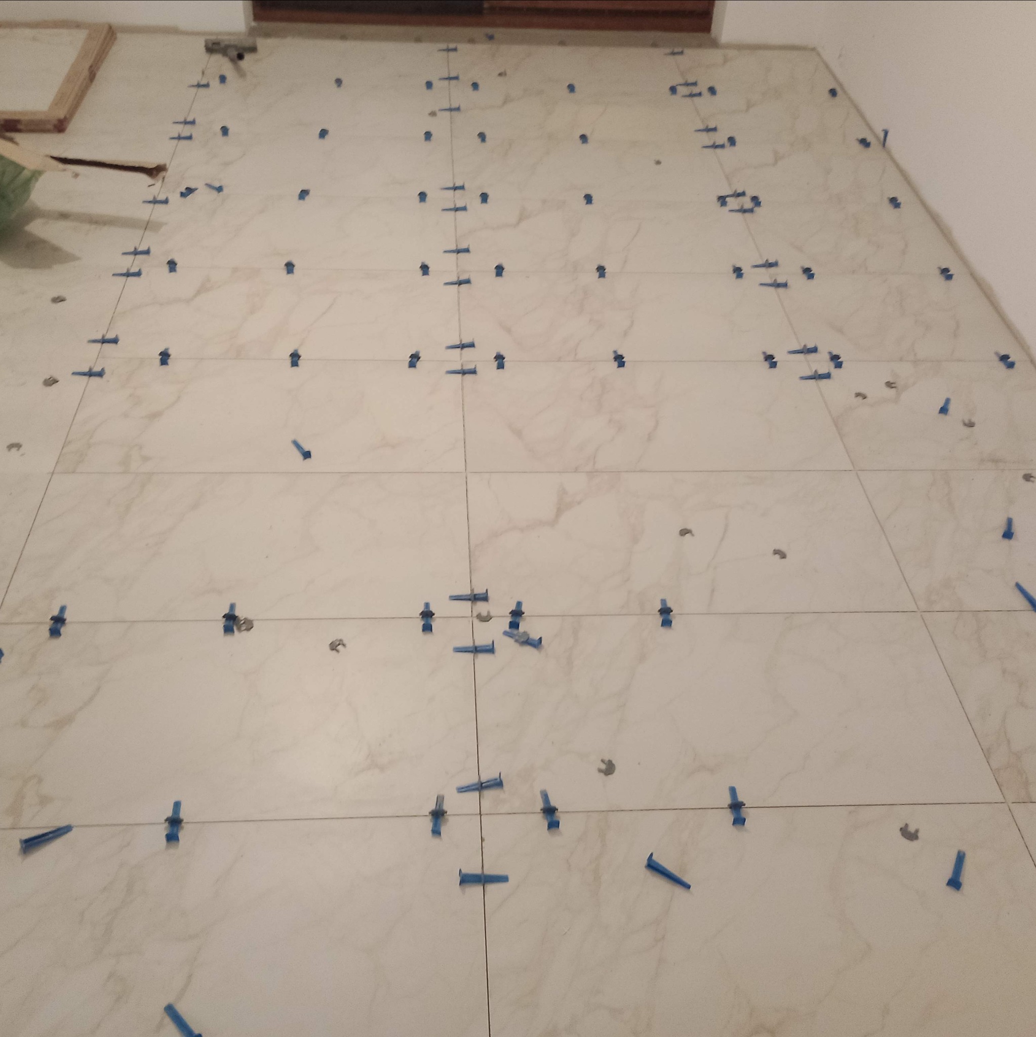 Układanie jasnych płytek podłogowych z systemem poziomowania, widoczne niebieskie kliny i klipsy dystansowe.